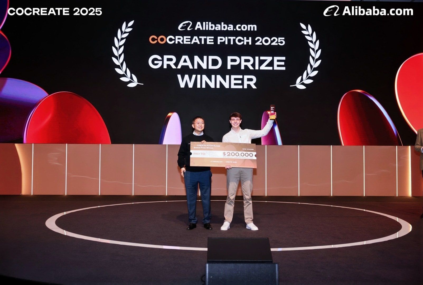 Harrison Nott on the CoCreate stage holding the $200,000 cheque and the trophy, under a screen reading Grand Prize Winner