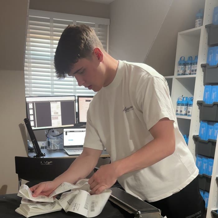 Harrison at a desk running a thermal label printer, shelves of stock behind him